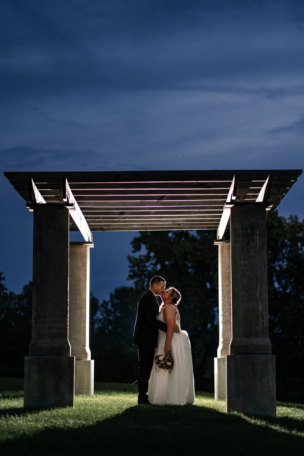 backlit image at dusk of bride and groom kissing under arbor 