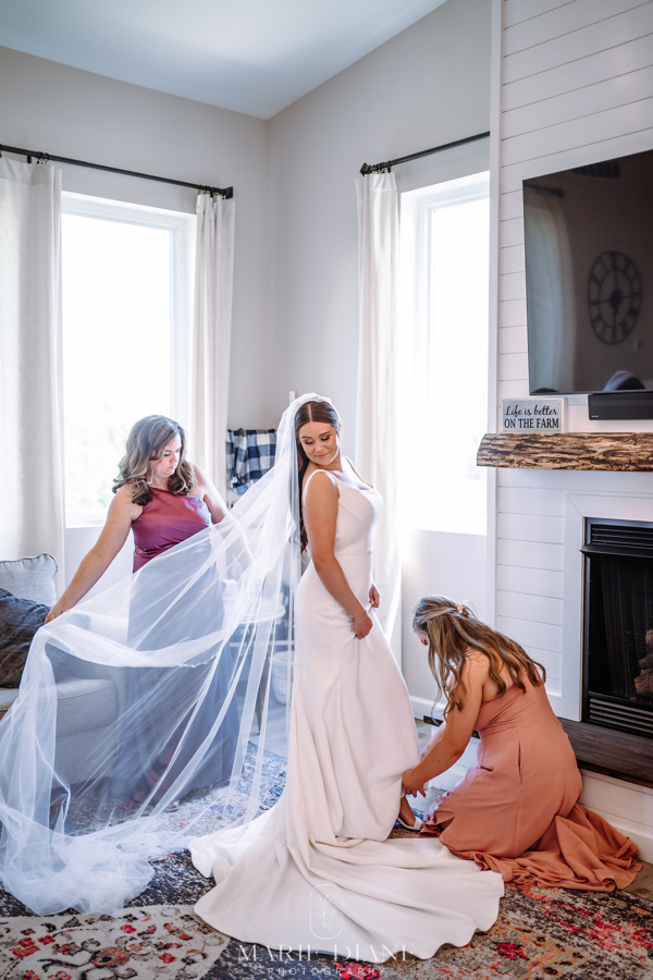 Mom of bride fixing veil on bride while maid of honor adjusts bride's shoe strap
